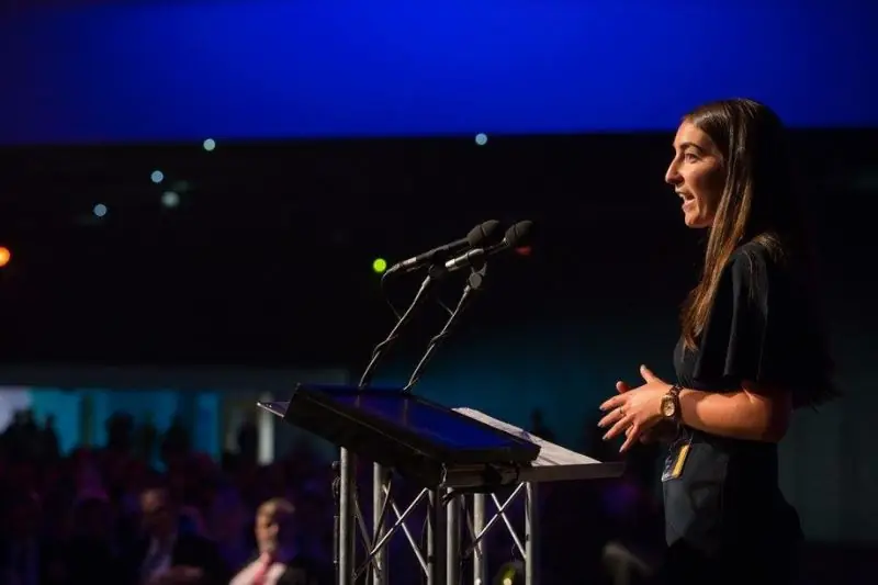 Speaking at a lectern on a darkened stage