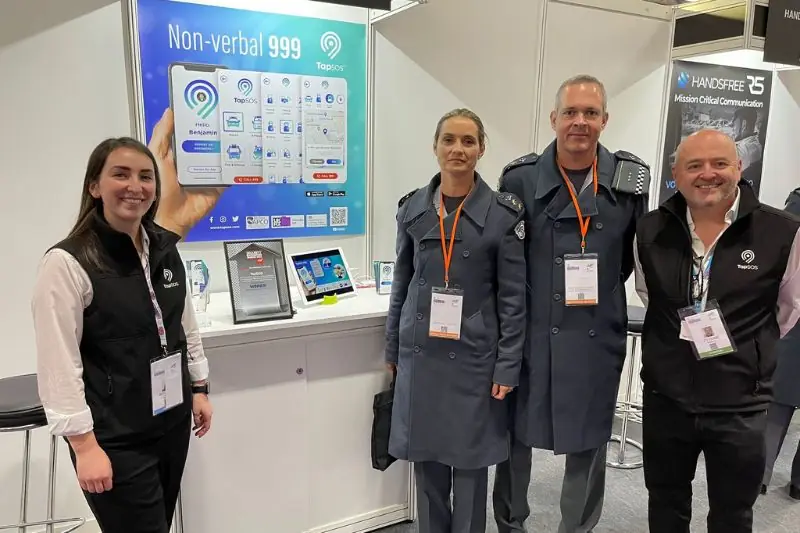 TapSOS exhibition stand with a large Non-verbal 999 banner, with the team and two uniformed visitors